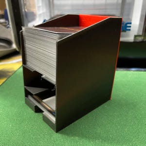 May include: A black card holder with a red interior. The holder is divided into sections, with a large stack of cards visible in the top compartment. The holder is made of a rigid material and sits on a green surface.