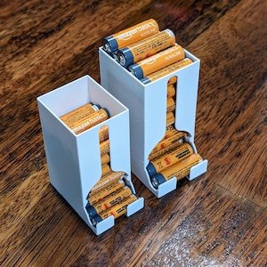 Puede incluir: Dos organizadores de baterías blancos impresos en 3D llenos de pilas alcalinas Amazon Basics naranjas y negras. Un organizador contiene pilas AA y el otro pilas AAA. Los organizadores están sobre una superficie de madera.