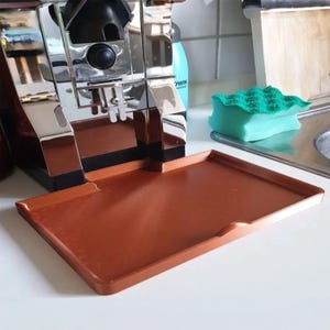 May include: A copper-coloured drip tray in front of a chrome espresso machine. The tray is rectangular with a raised edge. A green and blue sponge is in the background. The machine has a black base and a silver top.