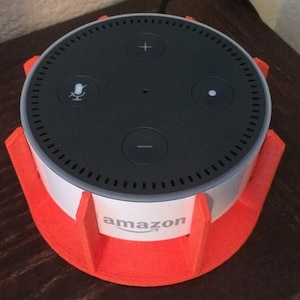 May include: A black and white Amazon smart speaker with a white band and the Amazon logo, set in a bright orange stand. The speaker has control buttons and a microphone icon on top. The stand is a vibrant, eye-catching accessory.
