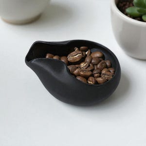 May include: A matte black ceramic scoop filled with roasted coffee beans. The scoop has a unique, curved design with a spout. The coffee beans are a rich brown color. The scoop is on a white surface.