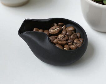 May include: A black ceramic scoop filled with roasted coffee beans. The scoop has a distinctive, curved design with a spout. The coffee beans are a rich brown colour. The scoop is on a white surface.