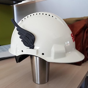 Op de afbeelding: Witte veiligheidshelm met een zwarte vleugel aan de zijkant. De helm heeft een gebogen klep en een reeks kleine ventilatiegaten aan de bovenkant. Het logo "S" en "BUILD" is zichtbaar aan de zijkant.