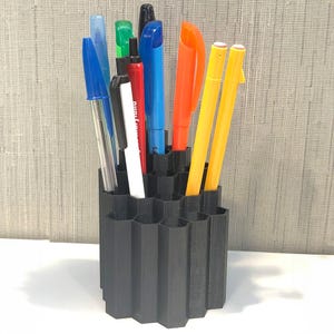 Peut inclure: Un porte-stylos noir hexagonal rempli de divers stylos et marqueurs. Les stylos sont de couleurs bleu, vert, rouge, orange et jaune. Le support est en matériau foncé et posé sur une surface blanche.