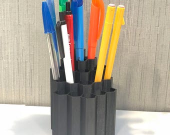 Op de afbeelding: Een zwarte, zeshoekige pennenhouder gevuld met diverse pennen en stiften. De pennen zijn in tinten blauw, groen, rood, oranje en geel. De houder is gemaakt van donker materiaal en staat op een wit oppervlak.