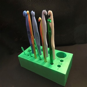 Peut inclure: Un support vert en plastique pour crochets, avec plusieurs trous et une variété de crochets colorés. Les crochets sont de différentes tailles et couleurs, dont le bleu, l'orange et le vert. Le support comporte des indications de taille.