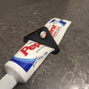 Könnte beinhalten: Ein schwarzer Zahnpasta-Tubenquetscher aus Kunststoff hält eine Zahnpastatube fest. Die Zahnpastatube ist weiß mit blauen Akzenten und dem Markennamen "Pep" in Rot. Die Tube ist mit "Whitens Teeth" und "NET WT. 156g" beschriftet.