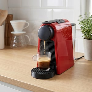Op de afbeelding: Een rode espressomachine met een zwarte boven- en onderkant, die koffie in een helder glazen kopje doseert. Stoom stijgt op van de vers gezette espresso. De machine staat op een licht houten aanrecht.
