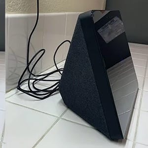 Peut inclure: Un appareil domestique intelligent triangulaire avec une base en tissu gris foncé et un écran noir. Un cordon d'alimentation noir est connecté à l'appareil. L'appareil est sur une surface carrelée blanche.