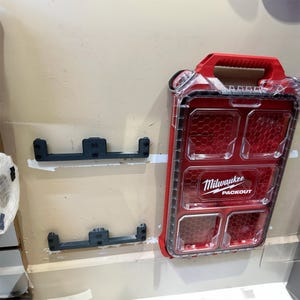 Peut inclure: Un conteneur de stockage Milwaukee Packout rouge et transparent avec une poignée rouge. Le conteneur présente un motif en nid d'abeille et le logo Milwaukee Packout. Deux supports de montage noirs sont également visibles.