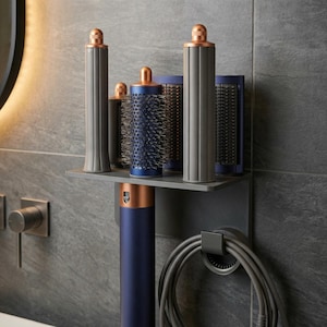 May include: A modern hair styling tool set in a bathroom setting. The set includes a dark blue and copper-colored hair dryer, various brush attachments, and a wall-mounted holder. The color scheme is primarily gray, blue, and copper.