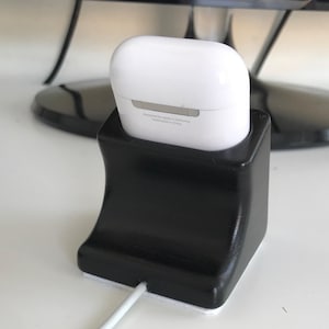 以下が含まれることがあります： 白いAirPodsケースを保持する、黒い湾曲した充電スタンド。スタンドは滑らかで光沢のある仕上げで、白い充電ケーブルがベースに接続されています。AirPodsケースは白で、シルバーのアクセントが付いています。