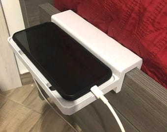 May include: A white plastic bedside shelf clamped onto a wooden headboard. The rectangular shelf features a recessed area holding a black smartphone connected to a white charging cable that extends downward from the device's bottom port.