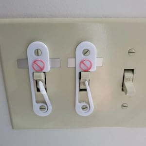 May include: White plastic light switch covers with a red prohibition symbol, installed on a beige wall plate. The covers are designed to prevent accidental activation of the light switches. One switch is in the off position.