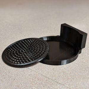 May include: A black, 3D-printed soap dish with a textured, oval-shaped lid and a round base. The lid has a pattern of small, zig-zag shaped holes. The base has a curved back and is open.