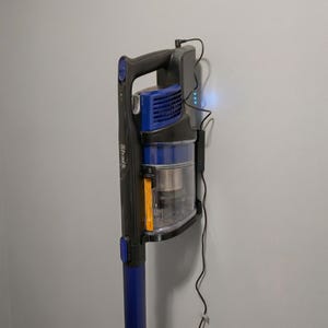 May include: A cordless Shark vacuum cleaner in a charging station. The vacuum has a black handle and body with blue accents. The dustbin is clear, and the charging station is mounted on a gray wall.
