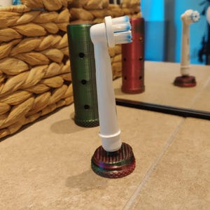 Puede incluir: Un cabezal de cepillo de dientes eléctrico blanco con cerdas azules se muestra sobre una base multicolor. Detrás del cepillo de dientes hay un soporte cilíndrico verde y rojo. El cepillo de dientes se refleja en un espejo.