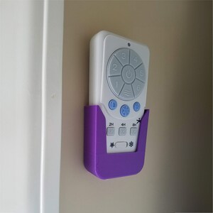 May include: A white ceiling fan remote control with gray buttons and a purple holder mounted on a beige wall. The remote has buttons for fan speed, a power button, and a timer. The holder is a solid purple color.