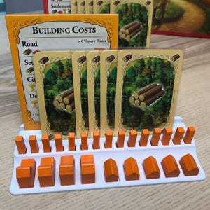 May include: A board game setup with cards depicting forests and logs, displayed in a white holder with orange wooden pieces. A card reads "BUILDING COSTS" and "Road". The game pieces are likely for a strategy game.