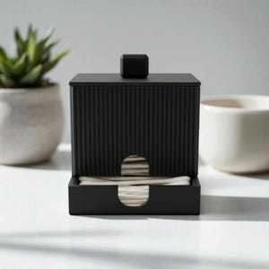 Può includere: Un dispenser nero rettangolare per cotton fioc con esterno a coste e un piccolo pomello quadrato sulla parte superiore. Il dispenser ha una fessura per un facile accesso ai cotton fioc. Una piccola pianta succulenta e una tazza di ceramica bianca sono sullo sfondo.