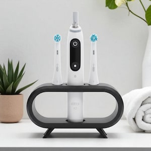 May include: A white and black electric toothbrush with two replacement brush heads, presented in a black oval-shaped holder. The brush heads have blue bristles. The toothbrush and holder are on a white surface.