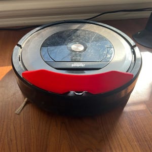 May include: A black and gray iRobot Roomba robotic vacuum cleaner with a red bumper. The vacuum is on a wooden floor. The Roomba has a circular design with a central control panel and a side brush.