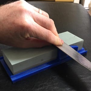 Puede incluir: Un cuchillo se afila sobre una piedra de afilar gris. La piedra de afilar se sujeta en un soporte de plástico azul. La hoja del cuchillo es plateada y refleja la luz. Se ve la mano de una persona.
