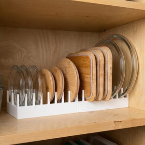 Könnte beinhalten: Ein weißer Kunststoff-Geschirr-Organizer hält eine Vielzahl von Glas- und Bambusdeckeln und Glasschalen. Die Deckel sind rund und quadratisch, mit einer hellbraunen Bambusoberfläche. Die Glasschalen sind klar und rund.