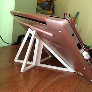 May include: A rose gold laptop rests on a white, geometric laptop stand. The stand features white, triangular supports. The laptop is angled upwards, displaying its design and ports.