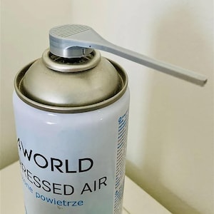Puede incluir: Una lata de aire comprimido con un diseño blanco y plateado. La lata tiene una boquilla plateada con una extensión larga y gris. El texto "WORLD PRESSED AIR" está impreso en la lata.