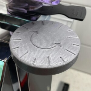 May include: A grey, circular dial with a textured surface and an arrow indicating clockwise rotation. The dial has evenly spaced lines around the edge, resembling a clock face. The object appears to be made of plastic.