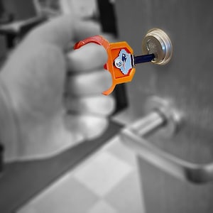 May include: An orange and red key holder with a blue and white emblem is inserted into a silver door lock. The key holder is designed to assist with turning the key. The background is blurred.