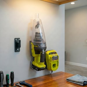 Op de afbeelding: Een draadloze handstofzuiger en oplader zijn aan een muur bevestigd. De stofzuiger is limoengroen en zwart met een doorzichtige stofbak. De oplader is geel en zwart. Een zwarte muurbevestiging bevindt zich links.