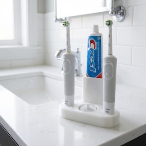 May include: Two white electric toothbrushes with green and white brush heads are in a white holder next to a tube of Crest toothpaste. The toothbrushes and toothpaste are on a white countertop in a bathroom.