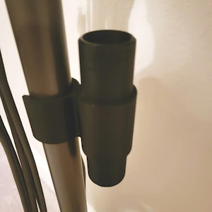 Puede incluir: Un accesorio negro para aspiradora, de forma cilíndrica con una base acampanada. El accesorio está conectado a un poste negro con una correa negra. El fondo es una pared blanca lisa.
