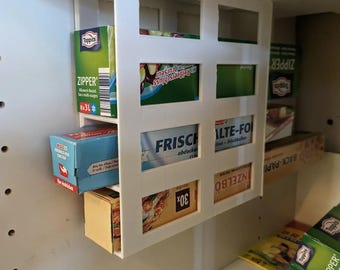 May include: A white, wall-mounted storage rack with multiple compartments, holding various boxes of food storage supplies. The boxes are green, blue, and yellow, with text labels. The rack is installed inside a cabinet.