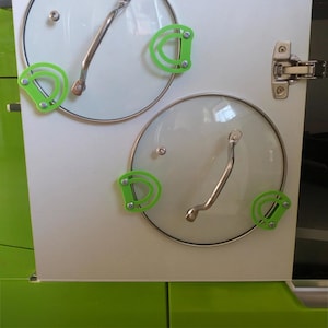 Peut inclure: Deux couvercles de casseroles en verre transparent avec des poignées en acier inoxydable et des supports de couvercle en silicone vert fixés à une porte d'armoire blanche. Les supports sont en forme de D et fixés avec de petites vis.