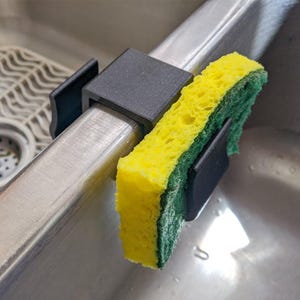May include: A black sponge holder attached to a stainless steel sink divider. A yellow and green sponge is held in place by the holder. The holder is made of a dark, matte material.