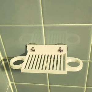 Puede incluir: Un organizador de ducha blanco, impreso en 3D, con un estante ranurado y dos soportes circulares. Tiene dos ventosas para montarlo en una pared alicatada. El organizador está diseñado para guardar artículos de ducha.