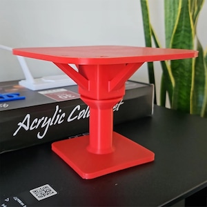 May include: A red 3D printed stand with a square base and a circular top. The stand is made of a smooth, glossy plastic.