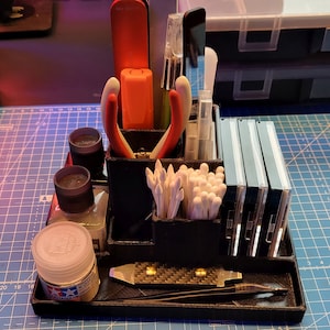 May include: A black organiser with multiple compartments for storing small items. It includes a pair of red pliers, a red marker, a set of cotton buds, and several small bottles of paint.