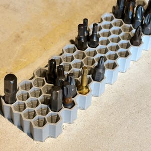 Op de afbeelding: Een lichtgrijze, honingraatvormige bithouder gevuld met diverse donker metalen en goudkleurige schroevendraaierbits. De houder is gemaakt van plastic en is ontworpen om verschillende soorten bits te organiseren en op te bergen.
