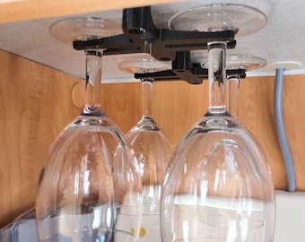 May include: A black wine glass rack mounted to a cabinet ceiling, holding four clear wine glasses. The rack has a suction cup design for easy installation. The glasses are suspended upside down, showcasing their elegant shape.