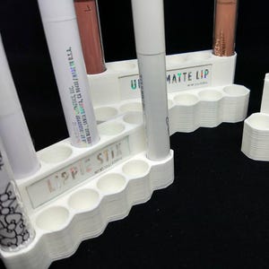 May include: A white 3D-printed lipstick organizer with multiple circular slots. The organizer holds various lip products, including lipsticks and lip glosses. The text "LIPPIE STIX" and "MATTE LIP" are visible on the organizer.