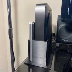 May include: A black and silver computer accessory holder. The holder is designed to hold a black computer device. The holder is made of a textured material and sits on a black surface. A black cable and a speaker are visible in the background.