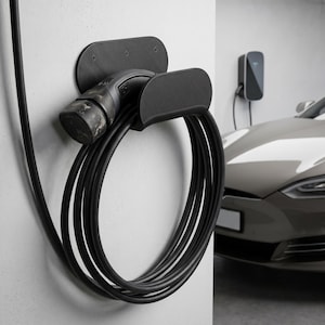 May include: A black electric vehicle charging station mounted on a white wall. The charging cable is coiled and held by a black bracket. A gray electric car is partially visible in the background.