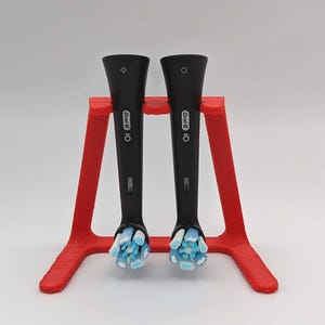 May include: Two black Oral-B iO toothbrush heads with blue and white bristles are held in a red 3D-printed stand. The stand has a simple design, and the toothbrush heads are positioned vertically.