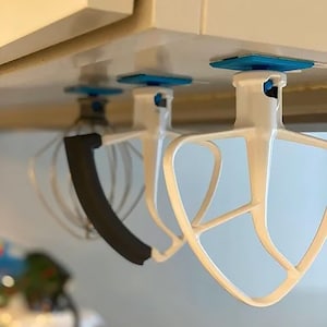 Könnte beinhalten: Drei weiße Küchenmixer-Aufsätze hängen kopfüber unter einem Schrank. Die Aufsätze umfassen einen Schneebesen mit schwarzem Gummigriff und zwei verschiedene Rührwerkstile. Jeder Aufsatz ist mit einer blauen quadratischen Halterung befestigt.
