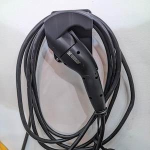 Puede incluir: Cargador de vehículo eléctrico negro con un cable largo y enrollado, montado en una pared blanca. El cargador se sujeta con un soporte negro. El mango del cargador tiene una serie de pequeños agujeros y una etiqueta con texto.
