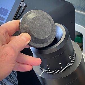 Peut inclure: Un couvercle gris foncé est soulevé d'un appareil cylindrique noir avec un cadran numéroté. Le cadran a des marques blanches et un pointeur blanc. L'appareil semble être un moulin à café ou un appareil de cuisine similaire.